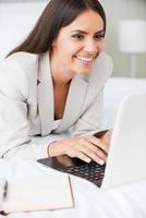 Staying in touch with colleagues in office. Confident young businesswoman in suit working on laptop and smiling while lying in bed at the hotel room photo