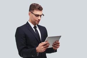 Concentrated at work. Handsome young man using digital tablet while standing against grey background photo