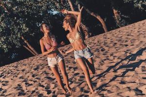 verano divertido. longitud total de dos mujeres jóvenes atractivas en pantalones cortos y traje de baño sonriendo mientras corre en la playa foto