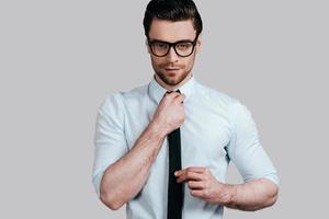 Perfect in his style.  Handsome young man in formalwear looking at camera and adjusting his tie while standing against grey background photo