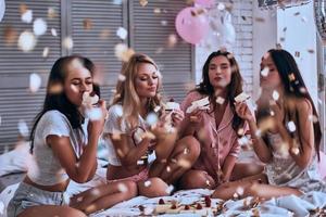 sabor increíble. cuatro hermosas mujeres jóvenes en pijama comiendo pastel mientras tienen una fiesta de pijamas en el dormitorio foto