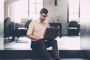 Man at work. Confident young man in smart casual wear working on laptop while sitting in office photo