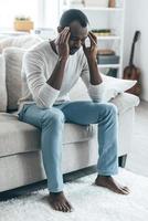 Stressed and tired. Frustrated young African man touching head with hands and keeping eyes closed while sitting on the sofa at home photo