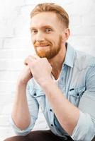 He is got a charming personality. Cheerful bearded man holding hands on chin and looking at camera while sitting against brick wall photo