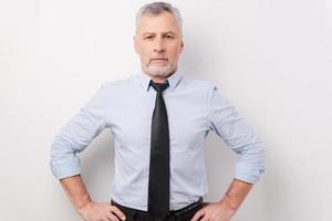 hombre de negocios seguro y exitoso. un anciano de pelo gris confiado con camisa y corbata tomándose de la mano en la cadera mientras se enfrenta a un fondo blanco foto