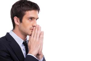 Begging for success. Side view of thoughtful young man in formalwear holding hands clasped near face and looking away while standing isolated on white background photo