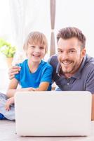Spending their free time together. Happy father embracing his cheerful little son while both sitting on the floor with laptop near them photo