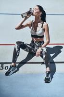 Time to rest. Top view of young woman in sports clothing drinking water while sitting on the running track outdoors photo