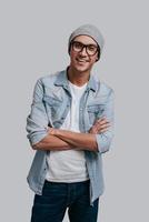 Young and carefree. Good looking young man in jeans shirt and headwear keeping arms crossed and looking at camera with smile while standing against grey background photo
