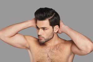 He is totally charming. Handsome young man looking away and keeping hands behind head while standing against grey background photo