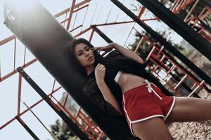 belleza confiada. bella joven con ropa deportiva ajustando su camiseta mientras se apoya en el saco de boxeo al aire libre foto