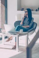 Lost in business thoughts. Thoughtful young beautiful woman holding hand on chin and looking through a window while sitting at her working place photo