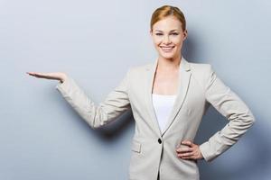 Your advertisement in her hand. Confident young businesswoman holding copy space and holding hand on hip while standing against grey background photo