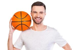 Listo para jugar. joven confiado que lleva una pelota de baloncesto en el hombro y sonríe mientras está de pie contra el fondo blanco foto