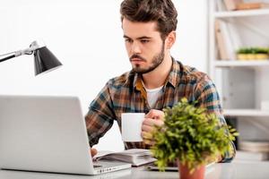 asegurándose de que toda la tarea se realice. un joven confiado que trabaja en una laptop y sostiene una taza de café mientras se sienta en su lugar de trabajo foto