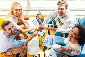 The best business team ever. Top view of group of smiling young people holding their hands clasped while sitting in the wooden desk together photo