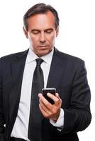 Checking business messages. Confident mature man in shirt and tie holding mobile phone and looking at it while standing isolated on white background photo