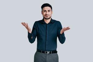 Confused young man shrugging shoulders and making face while standing against grey background photo