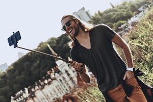 hora de hacerse un selfie. un joven apuesto con ropa informal sonriendo mientras se toma selfie al aire libre foto