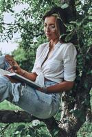 Perfect place to read. Attractive young woman in casual wear reading a book while sitting on the tree outdoors photo