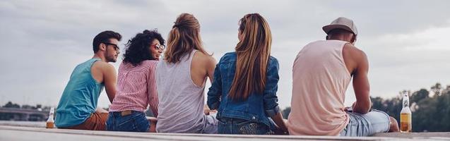 Young and carefree. Rear view of young people in casual wear smiling and talking while sitting on the pier photo