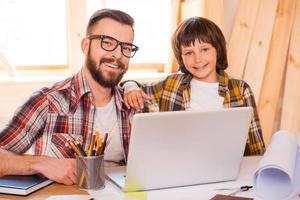 padre e hijo trabajando juntos. un joven alegre que trabaja en una laptop con su hijo mientras está sentado en el lugar de trabajo foto