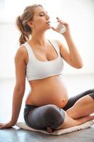 Only clean and fresh water. Portrait of pregnant young woman drinking water while sitting in lotus position photo