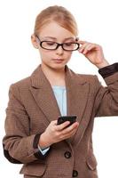 Reading an important message. Confident little girl in formalwear looking at the mobile phone and adjusting her glasses while standing isolated on white photo