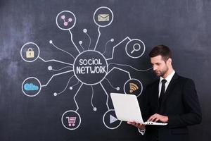 Social networking. Handsome young man working on laptop while standing against social network chalk drawing on blackboard photo