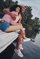 descansando juntos. feliz pareja joven abrazándose y sonriendo mientras se sienta en el muelle cerca del lago foto