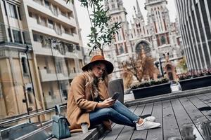 Little coffee break. Attractive young woman in hat and coat using her smart phone while sitting outdoors with church in the background photo