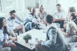 Welcome to team Group of confident business people sitting around the desk together while two men shaking hands and smiling photo