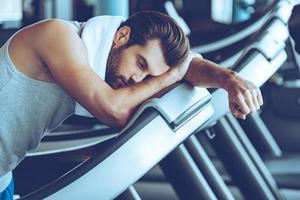 Cannot run anymore. Side view of young man in sportswear looking exhausted while leaning on treadmill at gym photo