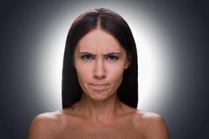 Woman grimacing. Portrait of young shirtless woman looking at camera and grimacing while standing against grey background photo