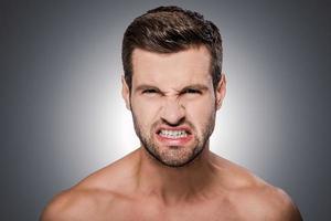 I am so angry Portrait of furious young shirtless man looking at camera and grimacing while standing against grey background photo