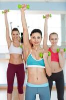 Entrenamiento con pesas. tres hermosas mujeres jóvenes con ropa deportiva haciendo ejercicio con pesas y sonriendo a la cámara foto