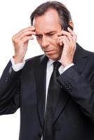 Bad news. Frustrated mature man in shirt and tie talking on the mobile phone and touching head with hand while standing isolated on white background photo