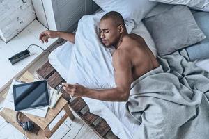 Time to wake up. Top view of young African man checking the time while lying in the bed at home photo