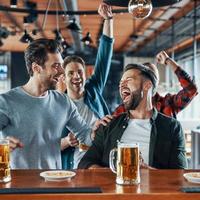Excited young men in casual clothing drinking beer and watching sport game while sitting in the pub photo