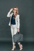 Young and smart. Full length of confident young woman in smart casual wear adjusting her eyeglasses and looking at camera while standing against grey background photo