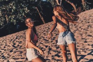 simplemente divirtiéndonos. dos atractivas mujeres jóvenes en pantalones cortos y trajes de baño sonriendo mientras corren en la playa foto