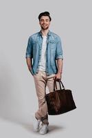 Stylish handsome man. Full length of good looking young man in casual wear carrying brown leather bag and smiling while standing against grey background photo