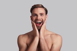 What a surprise Happy young man looking at camera and keeping head in hands while standing against grey background photo