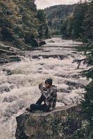 capturando recuerdos. apuesto joven moderno fotografiando mientras se sienta en la roca cerca del río foto