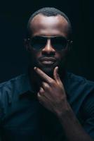 Trendy look. Portrait of handsome young African man in sunglasses holding hand on chin and looking at camera while being in front of black background photo