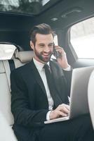 Great solutions every day. Handsome young businessman working on his laptop and talking on the phone while sitting in the car photo