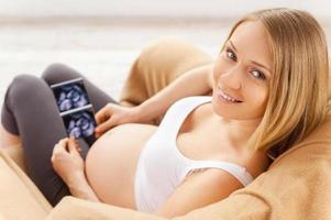 The first photo of my baby. Top view of happy pregnant woman sitting on the chair and holding x-ray image of her baby