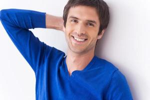 Handsome and self-confident. Handsome young man in blue sweater looking at camera and smiling while holding hand behind head and standing against grey background photo