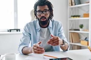Confident young African man in eyewear telling something and looking at camera while working indoors photo
