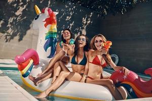 mujeres jóvenes atractivas en traje de baño sonriendo y apuntándote con pistolas de agua mientras flotan en un unicornio inflable en la piscina al aire libre foto